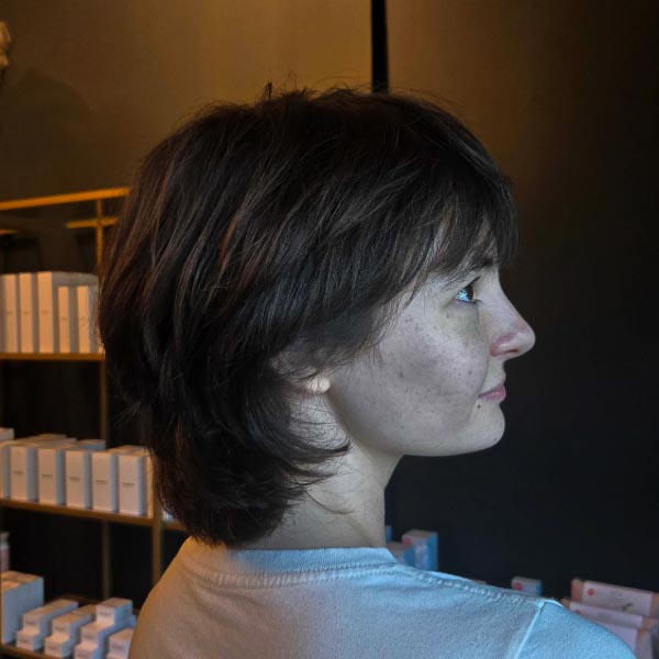 A person with short, layered brown hair is shown in profile, standing indoors against a dark wall with shelves lined with neatly arranged boxes in the background.