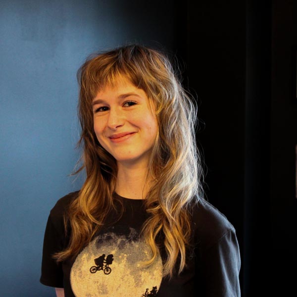 A person with wavy, light brown hair smiles softly while standing indoors against a dark wall. They are wearing a black T-shirt with a moon and bicycle graphic on the front.