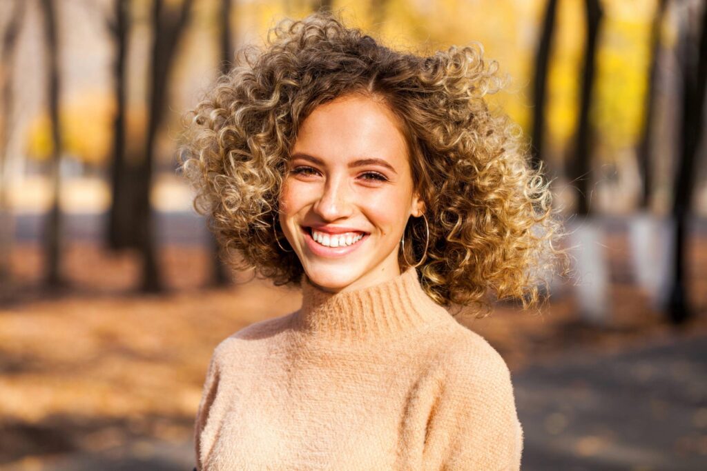A young woman with curly blonde hair and a beige sweater smiles outdoors in a park, her hair styling perfectly framed by autumn leaves and sunlight that highlights her face and hair.