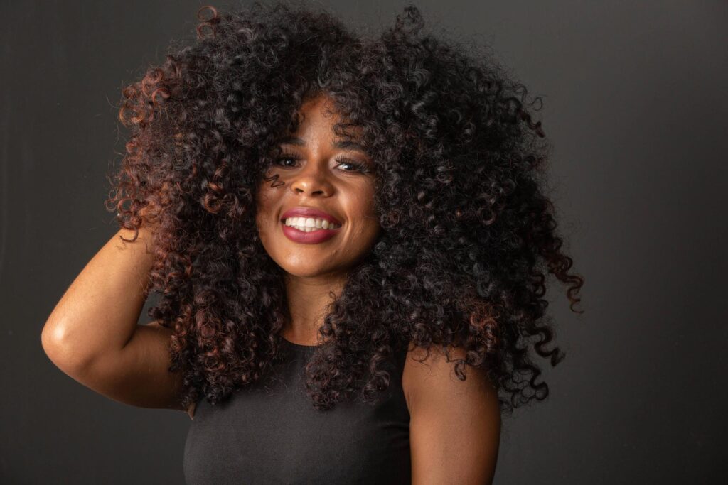 A woman with voluminous curls smiles brightly, wearing a black sleeveless top and posing against a plain dark background. Her hand is lifted to her hair, embodying the confidence of a curly hair stylist.