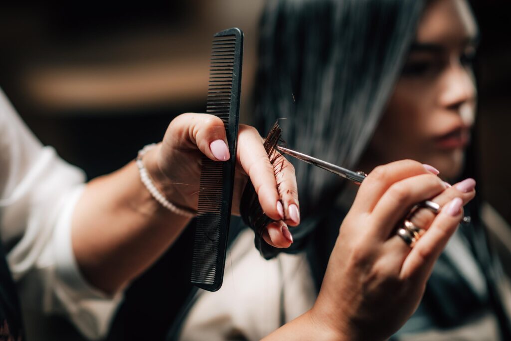 A close-up of a hairdresser’s hands expertly delivering precision haircuts, cutting a client's dark hair with scissors and a comb, carefully sectioning each strand between their fingers. The client is blurred in the background.
