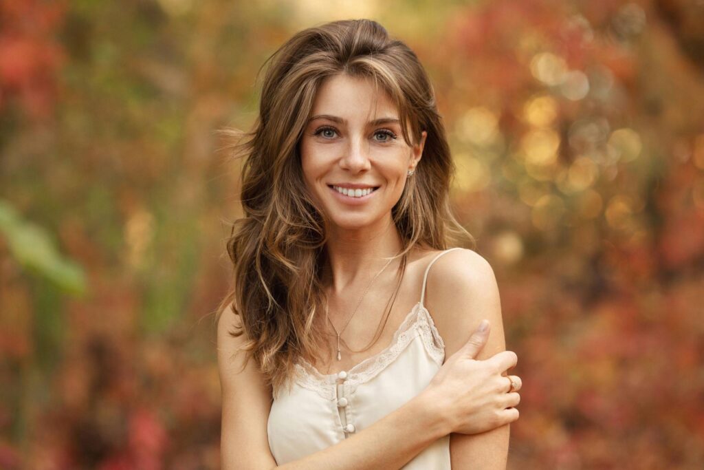 A young woman with long brown hair and a light camisole smiles at the camera, standing outdoors in front of a blurred background with lived in color from vibrant autumn foliage.