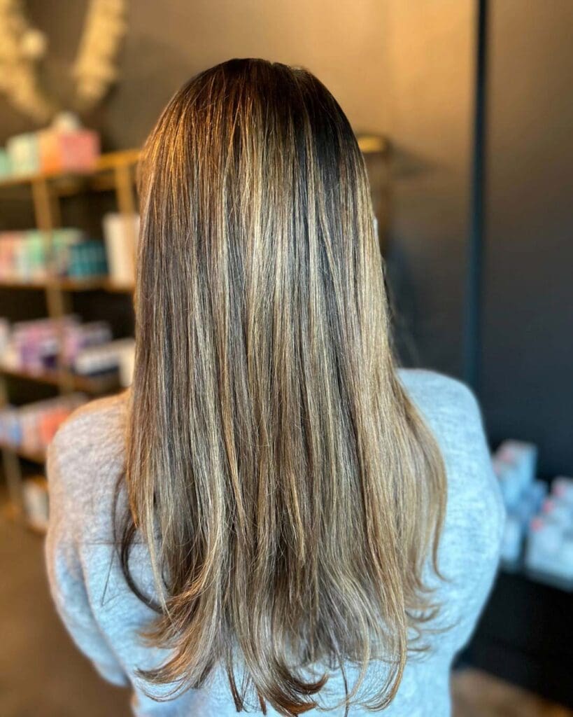 A client with long, straight hair showcasing fine golden blonde highlights and a dark base, styled in a polished, sleek blowout at Sunday Salon in Raleigh.