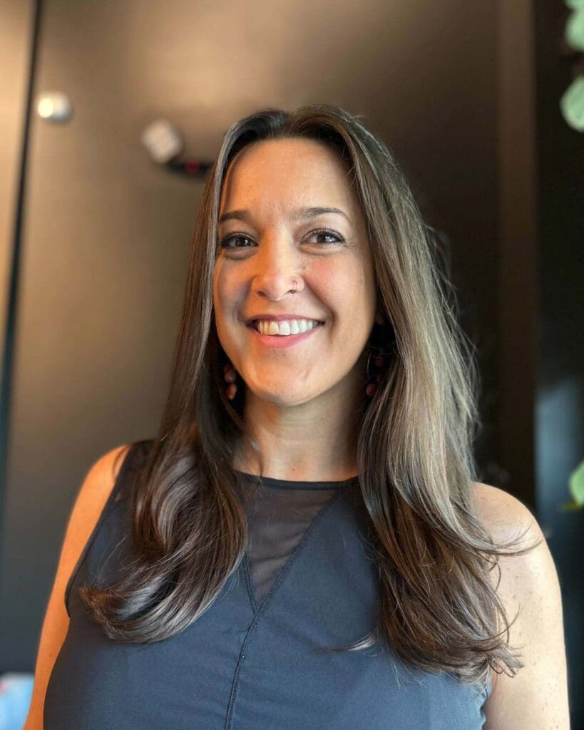 A smiling client showcasing a polished, long brunette blowout with subtle ash face-framing dimension and softly curled ends, created at Sunday Salon in Raleigh.
