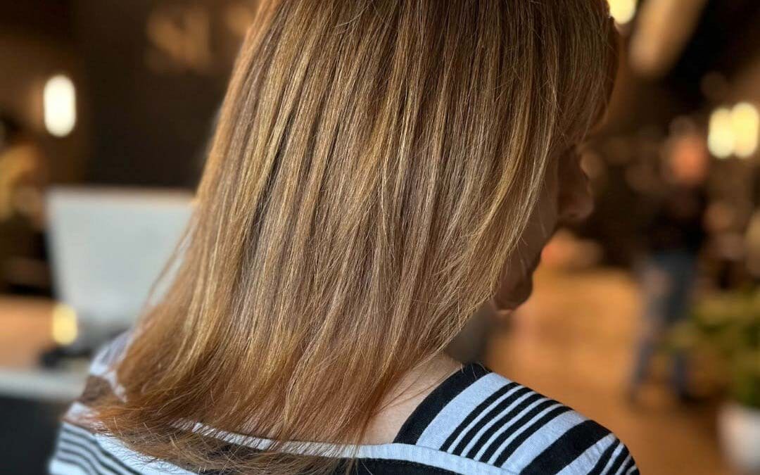 A client showcasing shoulder-length hair styled in a sleek and smooth blowout, featuring dimensional warm honey blonde highlights, created at Sunday Salon in Raleigh.