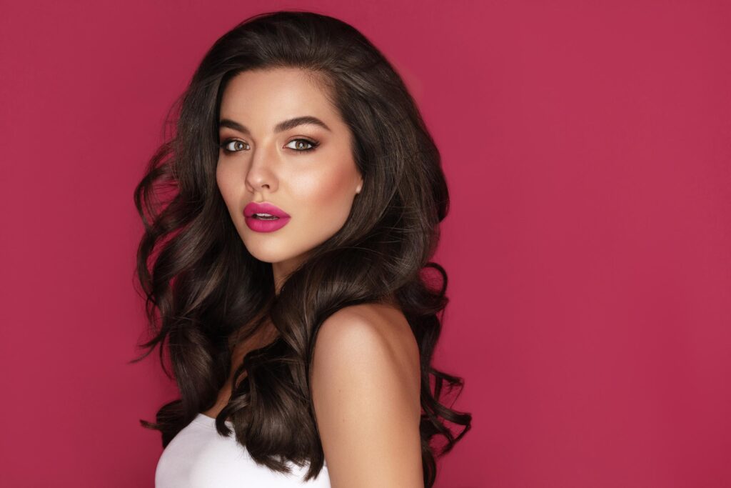 A woman with long, wavy brown hair styled to perfection, as if fresh from salon blowouts, and bold pink lipstick poses against a solid magenta background, wearing a white top and looking confidently at the camera.
