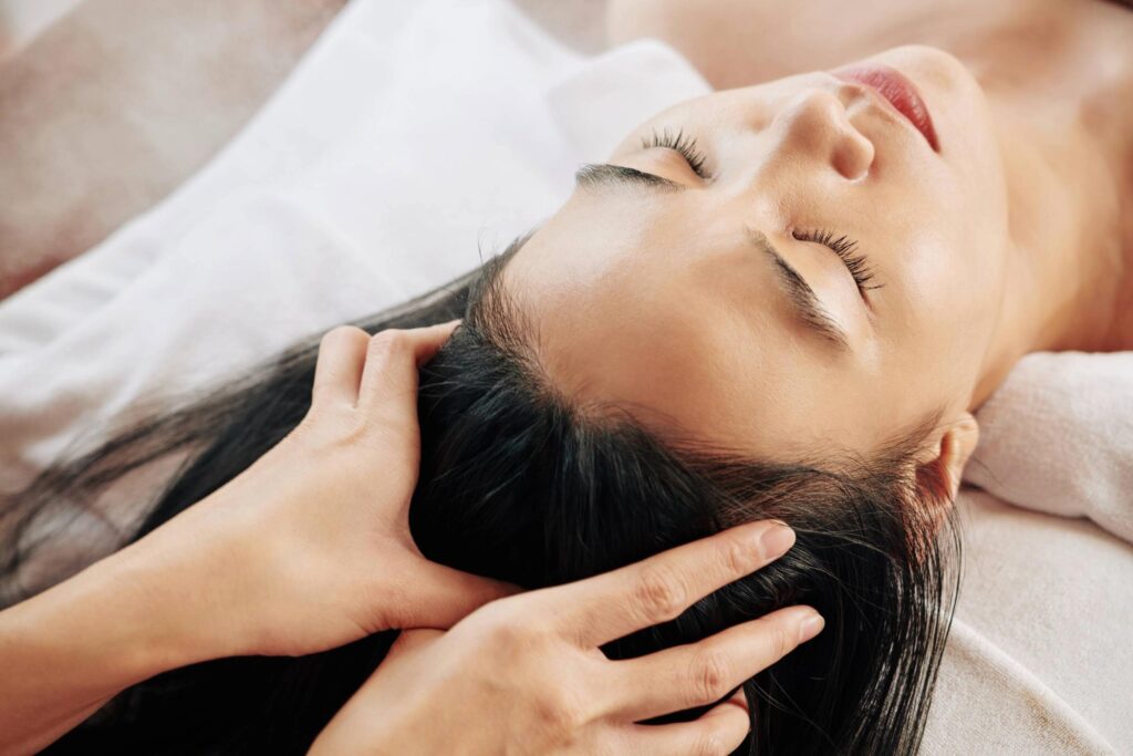 A person with closed eyes receives a relaxing scalp massage while lying on a white towel, conveying a sense of calm and wellness.
