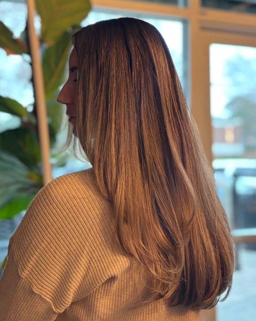 A side profile of a client showing off ultra-long, thick, warm caramel brunette hair styled in a sleek blowout with softly sweeping layered ends at Sunday Salon in Raleigh.