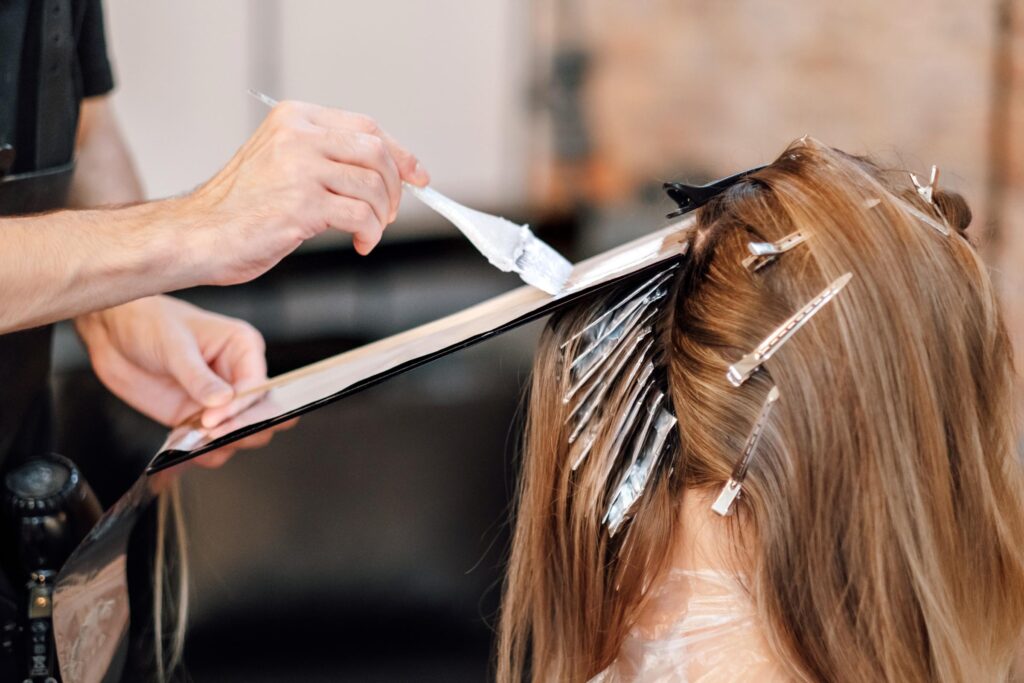 A hair colorist applies dye with a brush to a client's long hair, carefully sectioned with clips and foil for highlights. The client is wearing a protective cape.