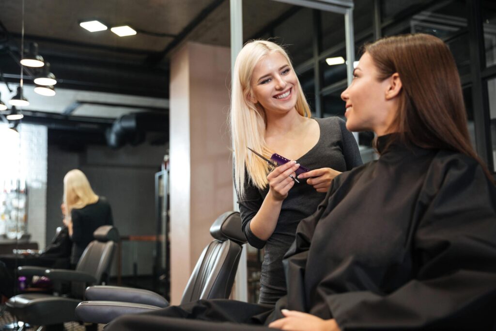 A professional hair stylist with blonde hair smiles while talking to a seated client with brown hair in a salon. The client is wearing a black salon cape, and the space features a modern, industrial interior.