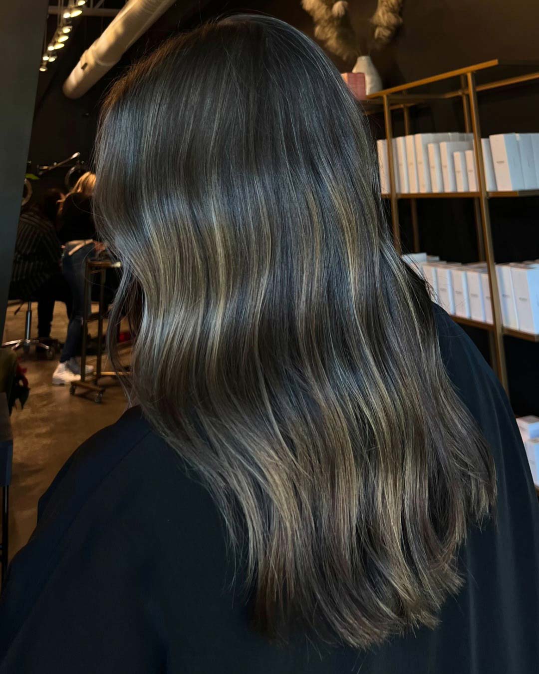 Delaney Neumann Stylist Sunday Salon Portfolio 1 A person with long, wavy, dark brown hair with subtle highlights sits in a modern salon, facing away from the camera. Shelves with white boxes and another person are visible in the softly lit background.