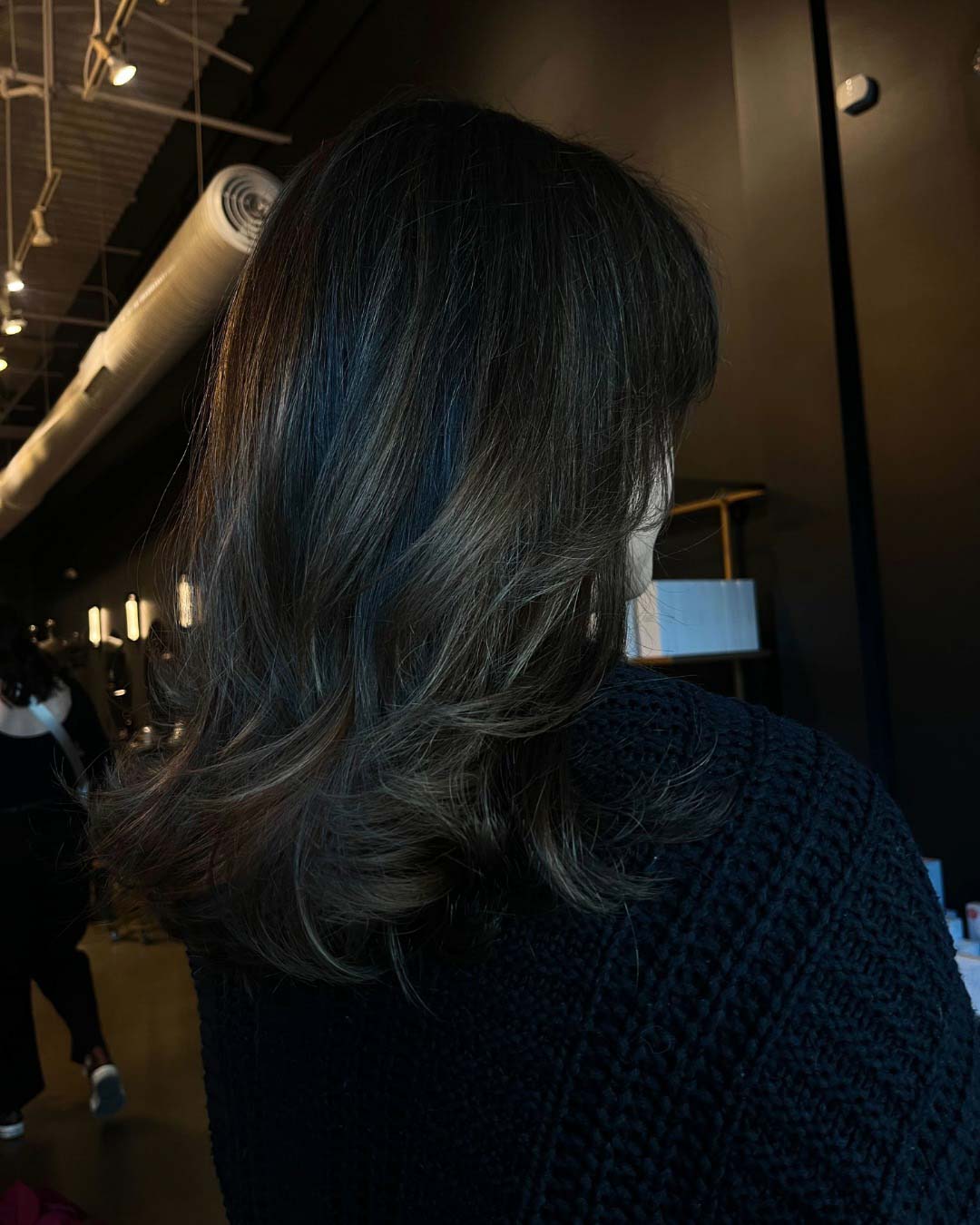 Delaney Neumann Stylist Sunday Salon Portfolio 2 A person with shoulder-length, dark brown wavy hair wears a textured black sweater, standing indoors with warm lighting and industrial decor in the background.