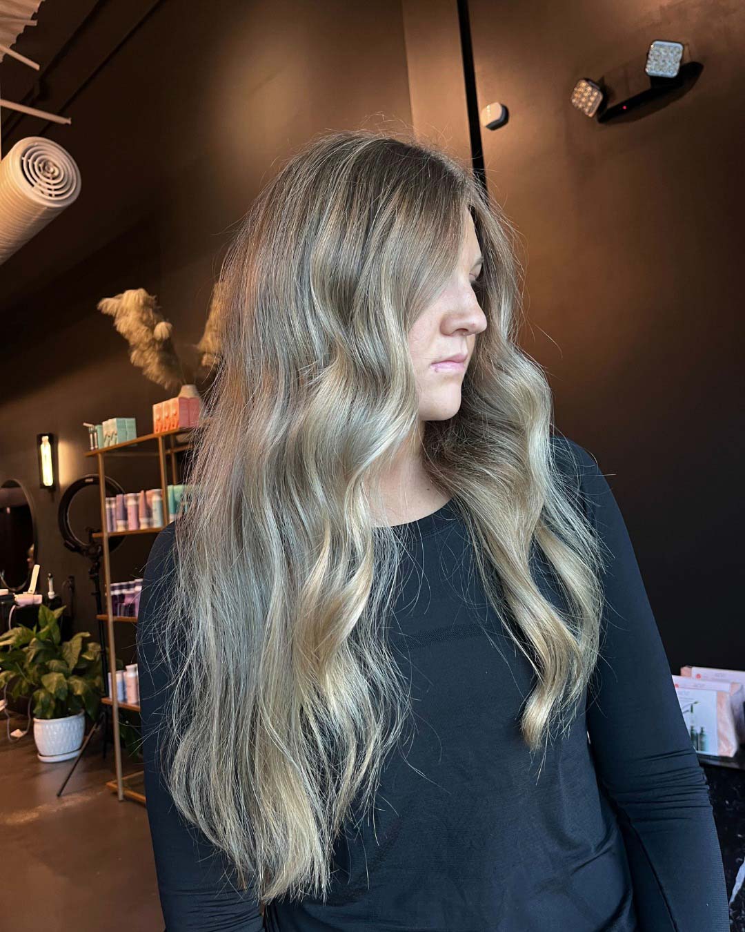 Delaney Neumann Stylist Sunday Salon Portfolio 3 A woman with long, wavy, ash-blonde hair wearing a black top stands in a modern salon, looking to the side. Shelves with hair products and plants are visible in the background.