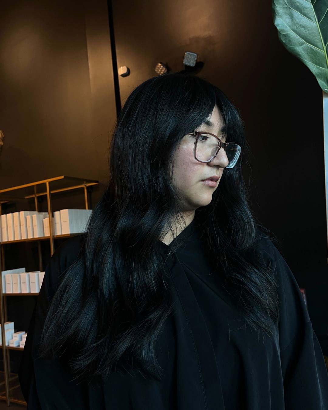 Delaney Neumann Stylist Sunday Salon Portfolio 4 A person with long, wavy black hair and glasses looks to the side, standing indoors against a dark background with shelves and a large green leaf visible. They are wearing a dark top.