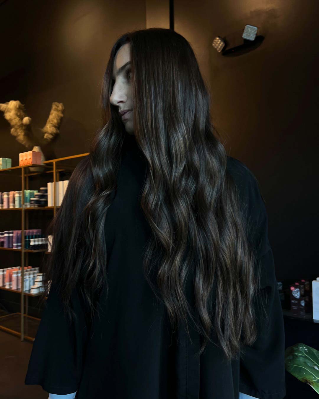 Delaney Neumann Stylist Sunday Salon Portfolio 5 A person with long, wavy, brunette hair wearing a black top stands indoors, looking to the side. Shelves with hair products and plants are visible in the background against dark walls.