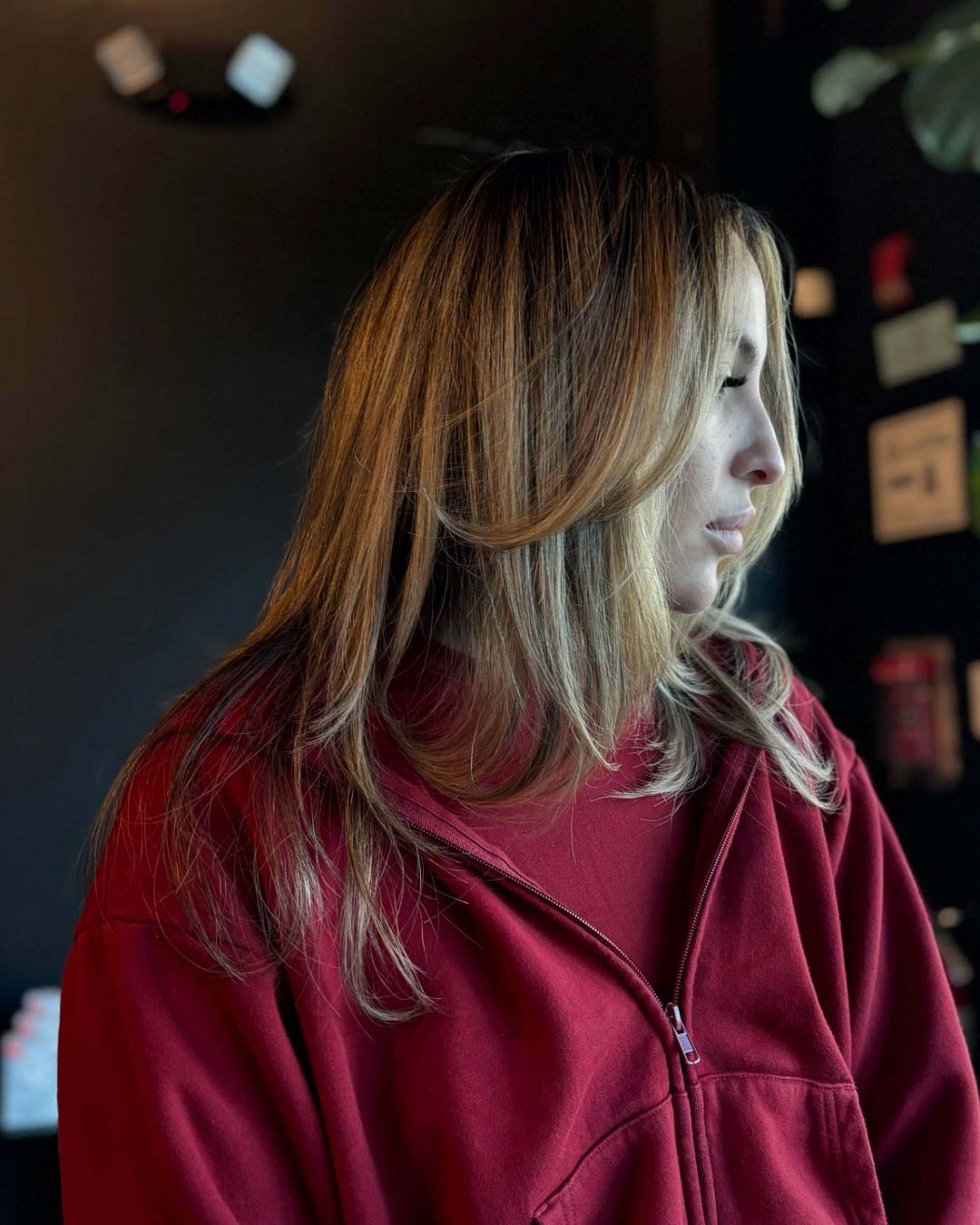 Jada_Handy_Portfolio_6 A woman with long, light brown hair wearing a red zip-up jacket looks to her right, standing indoors against a dark, softly lit background.