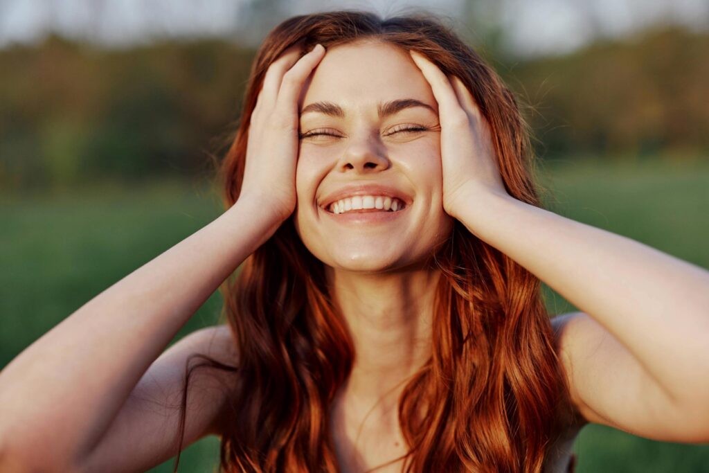 A young woman with vibrant red hair smiles brightly with her eyes closed, holding both sides of her face in delight. She is outdoors, framed by a blurred green background.