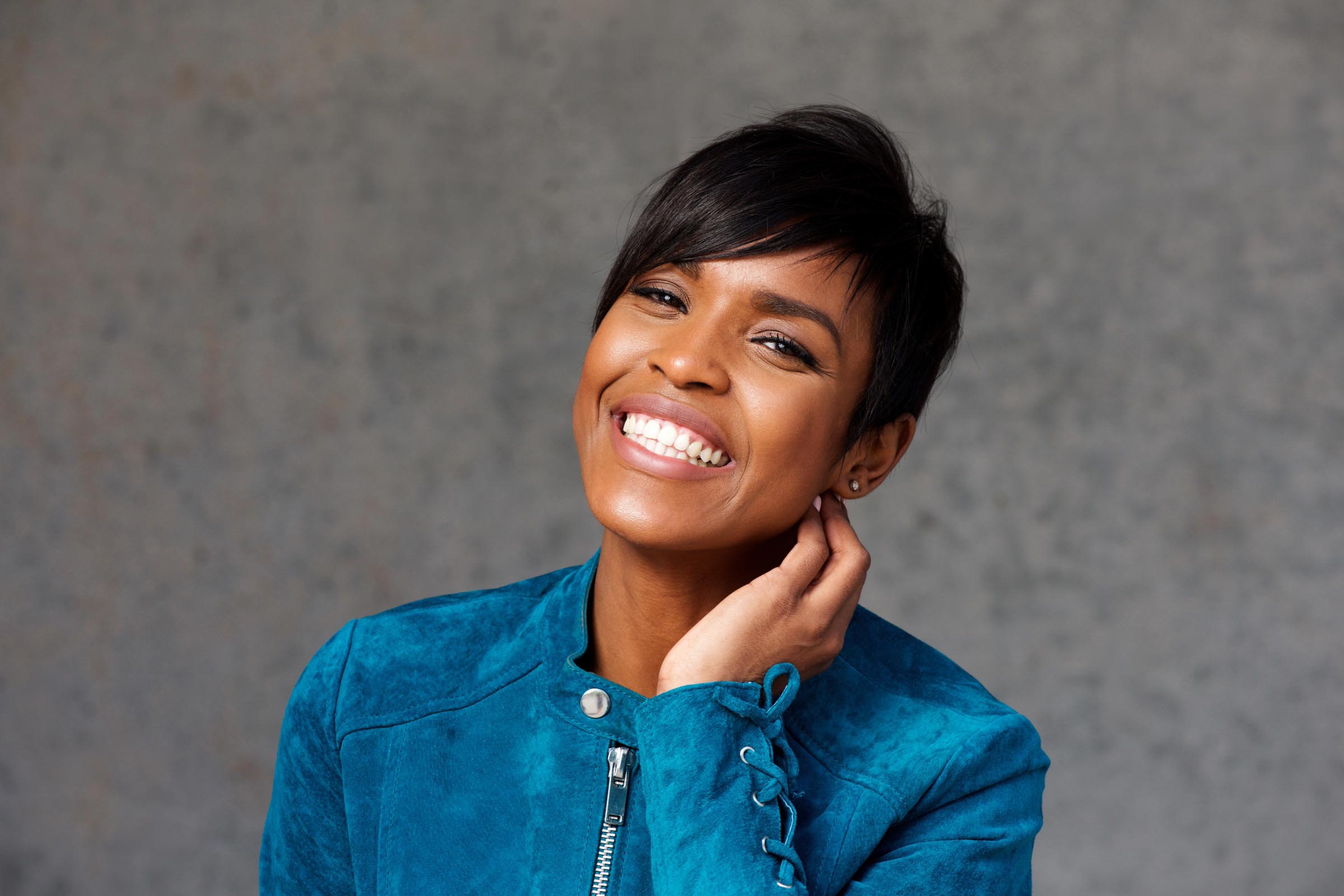 A woman with low maintenance short haircuts smiles brightly, tilting her head and touching her neck. She is wearing a teal suede jacket and is posed in front of a plain gray background.