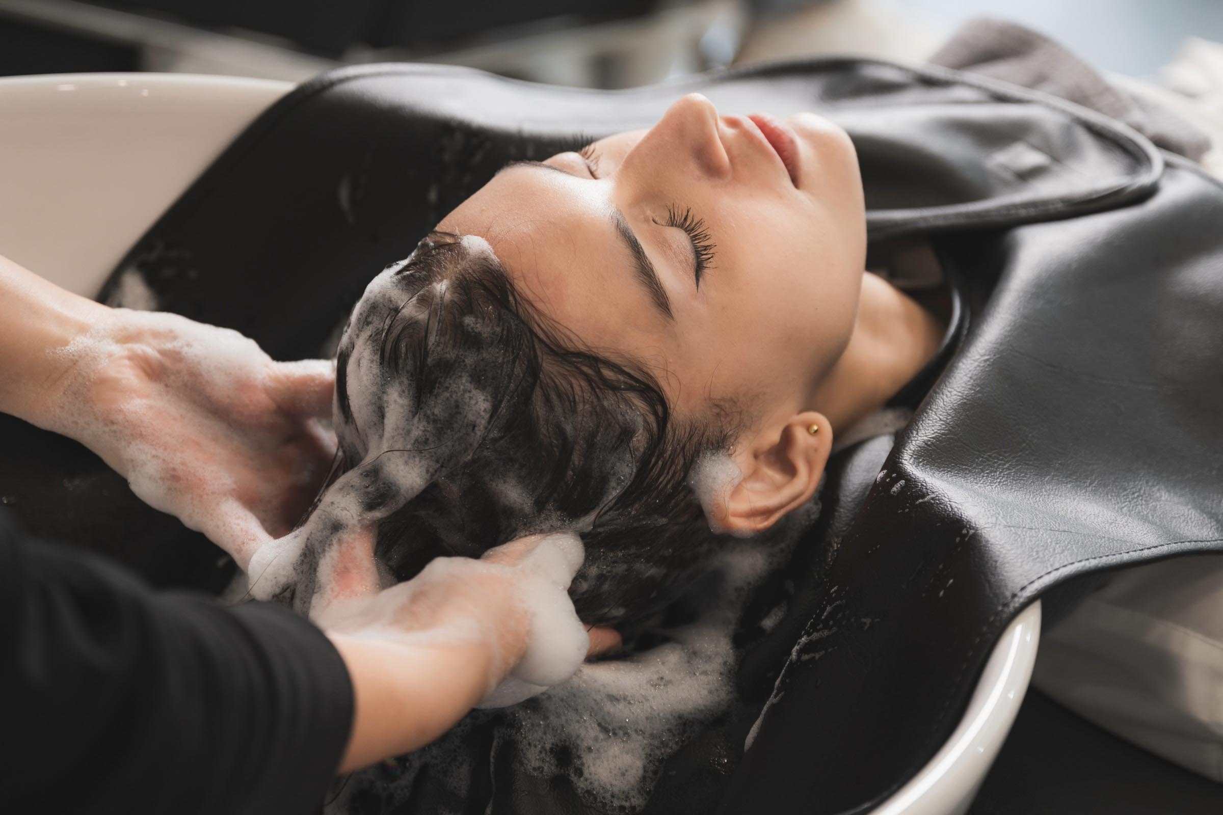 A person with closed eyes is reclining at a salon sink while someone washes their hair with foamy shampoo, enjoying relaxing Cary hair salon treatments.