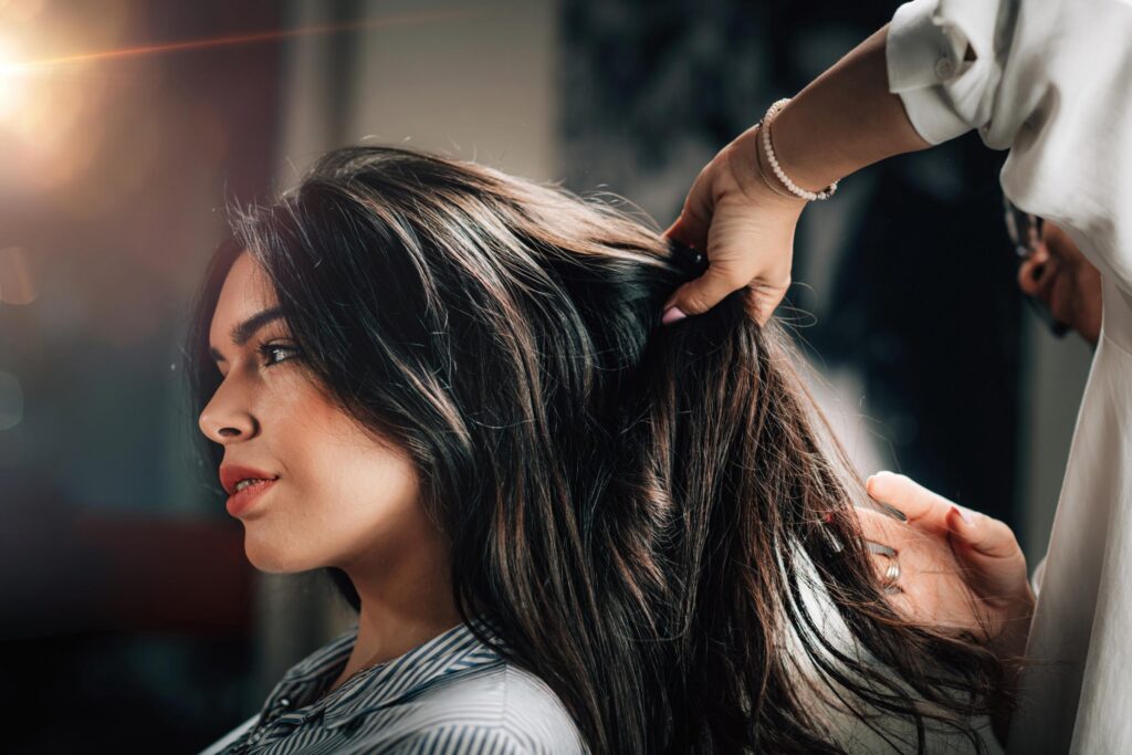 A woman with long, dark hair sits in a salon chair, while a stylist lifts sections of her hair. Warm light highlights the scene, creating a soft, professional salon atmosphere.