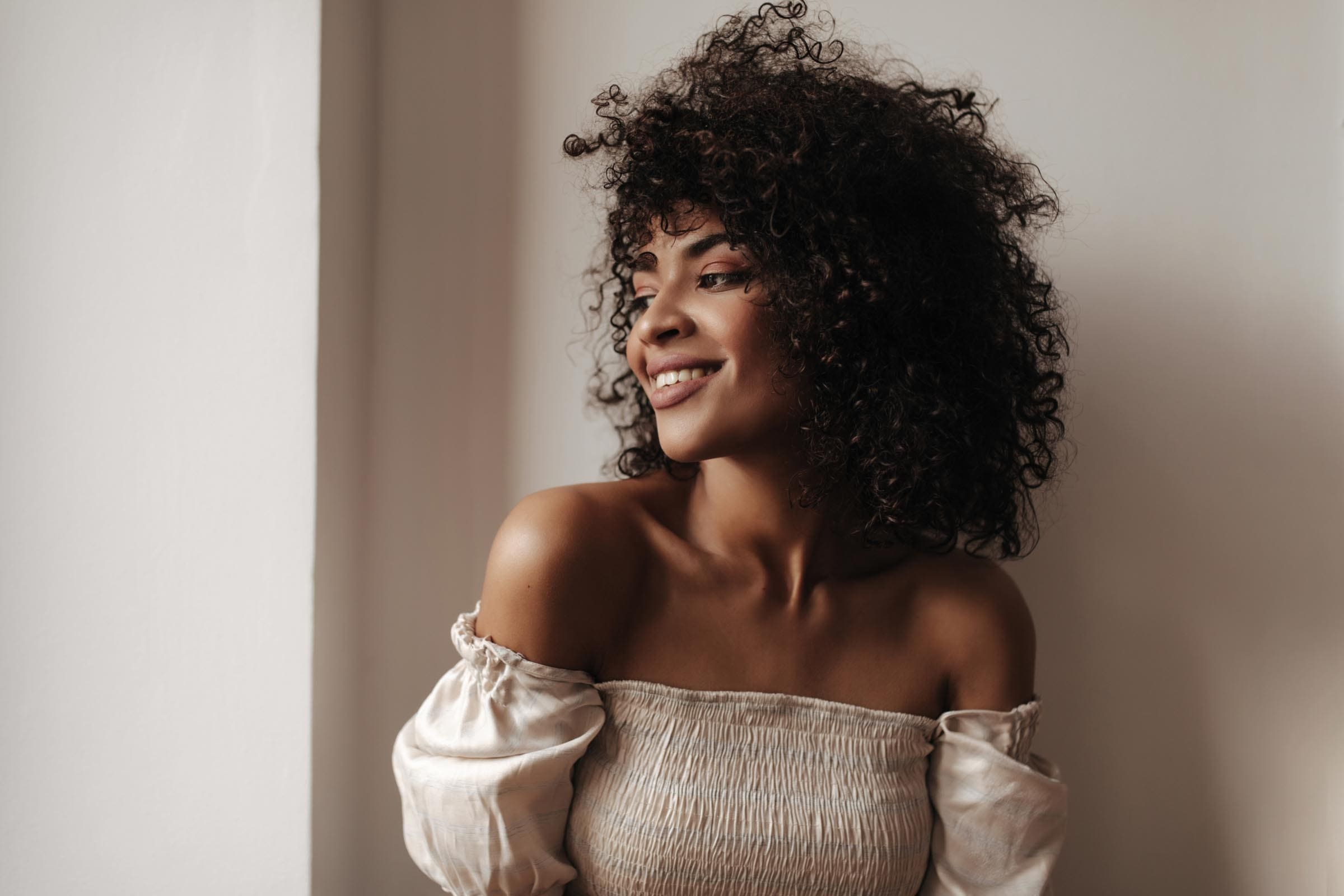 A woman with curly hair styled by a talented curly hair stylist wears an off-shoulder cream top and smiles while looking to the side, standing against a plain white wall in soft natural light.
