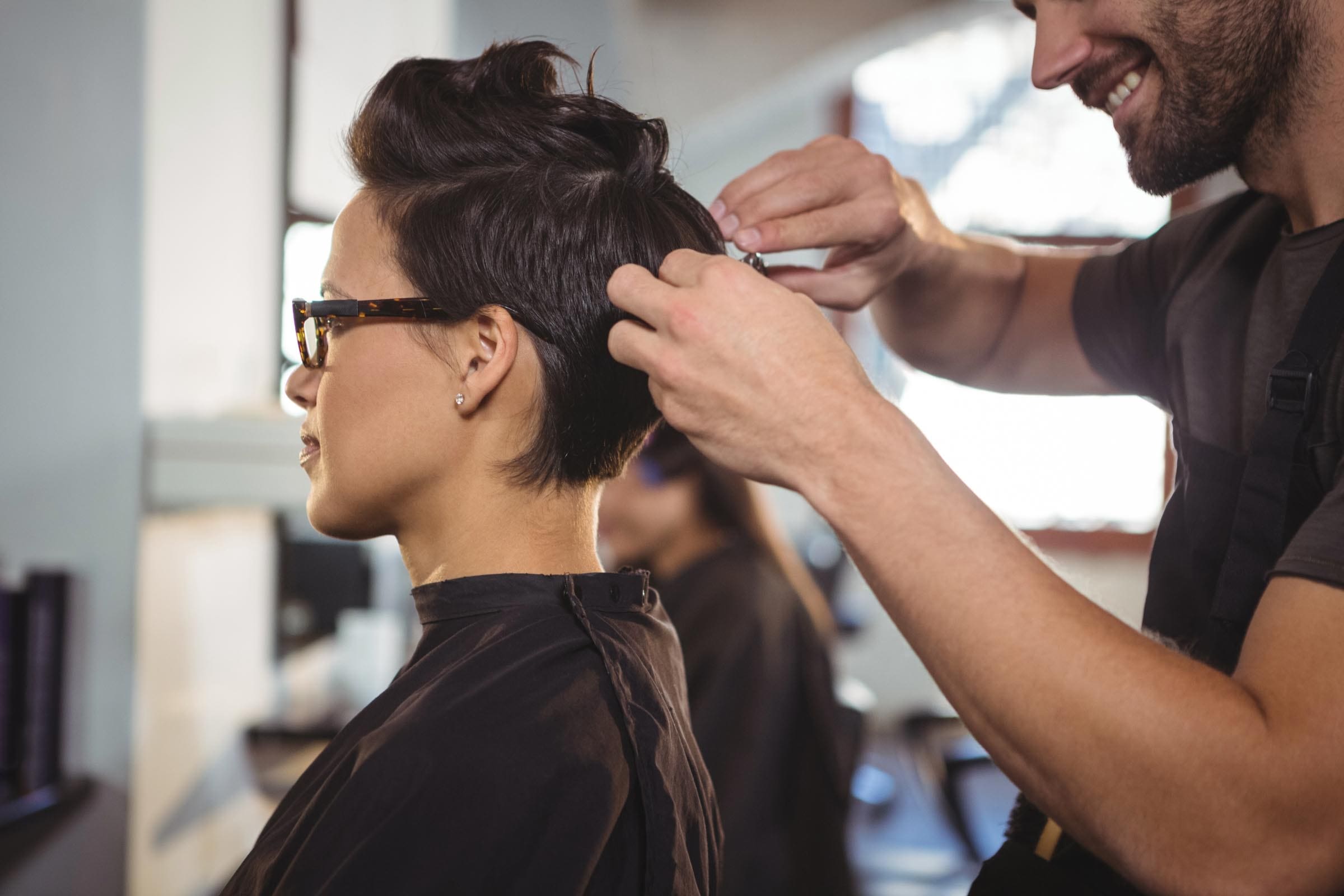 A hairstylist is cutting the short, dark hair of a person wearing glasses and a black cape in a brightly lit salon. After their hair consultation, the person smiles slightly as the stylist works with scissors.