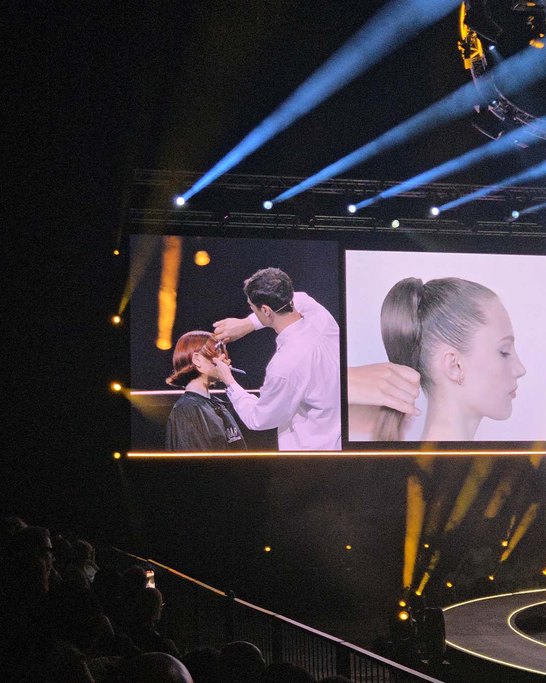 davines-euope-4 A stylist demonstrates a haircut and a ponytail hairstyle on a large projection screen above an audience.